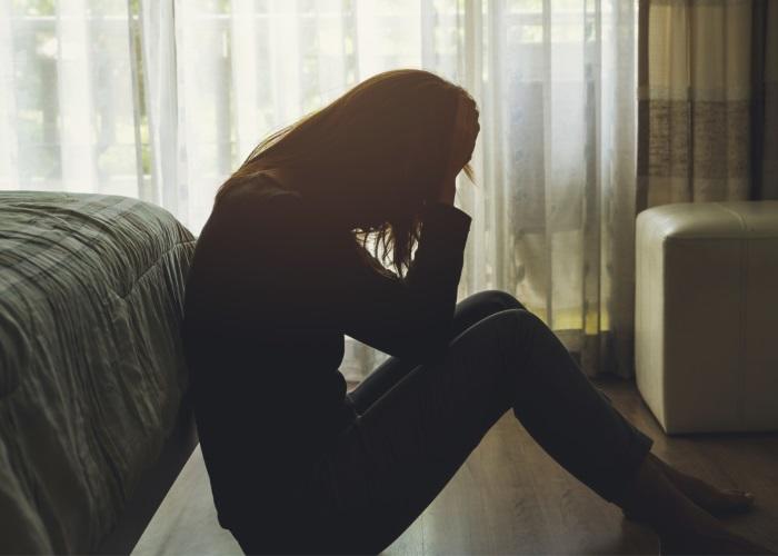 Frau sitzt in einem abgedunkelten Zimmer auf dem Boden und hält sich die Hände wegen einer Migräne-Attacke an den Kopf.