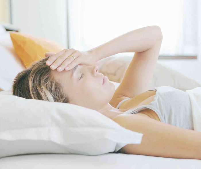 Frau liegt im Bett und hält sich den Kopf aufgrund ihrer hemiplegischen Migräne. Lähmungen sind weitere Symptome.
