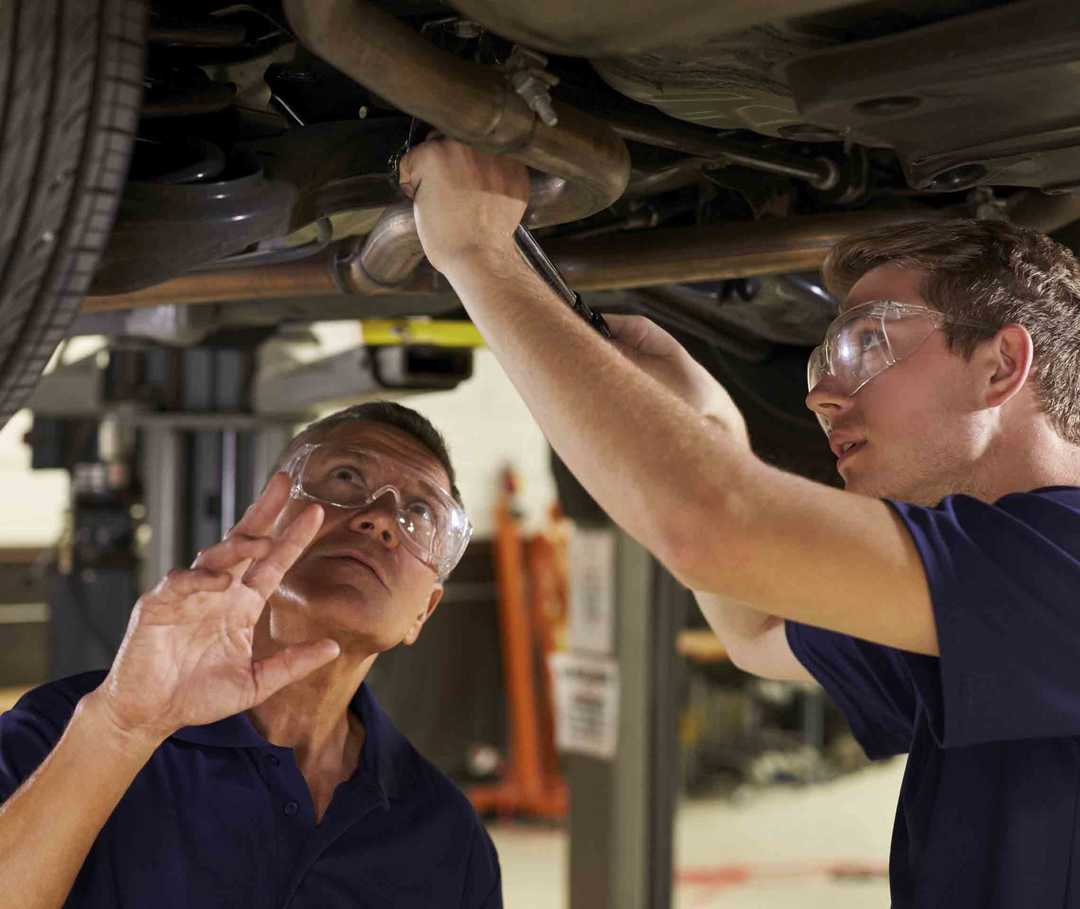Zwei Männer reparieren ein Auto in der Werkstatt: Einer leidet an Migräne, die ihn zwingt, kurzfristig seine Arbeit zu unterbrechen.