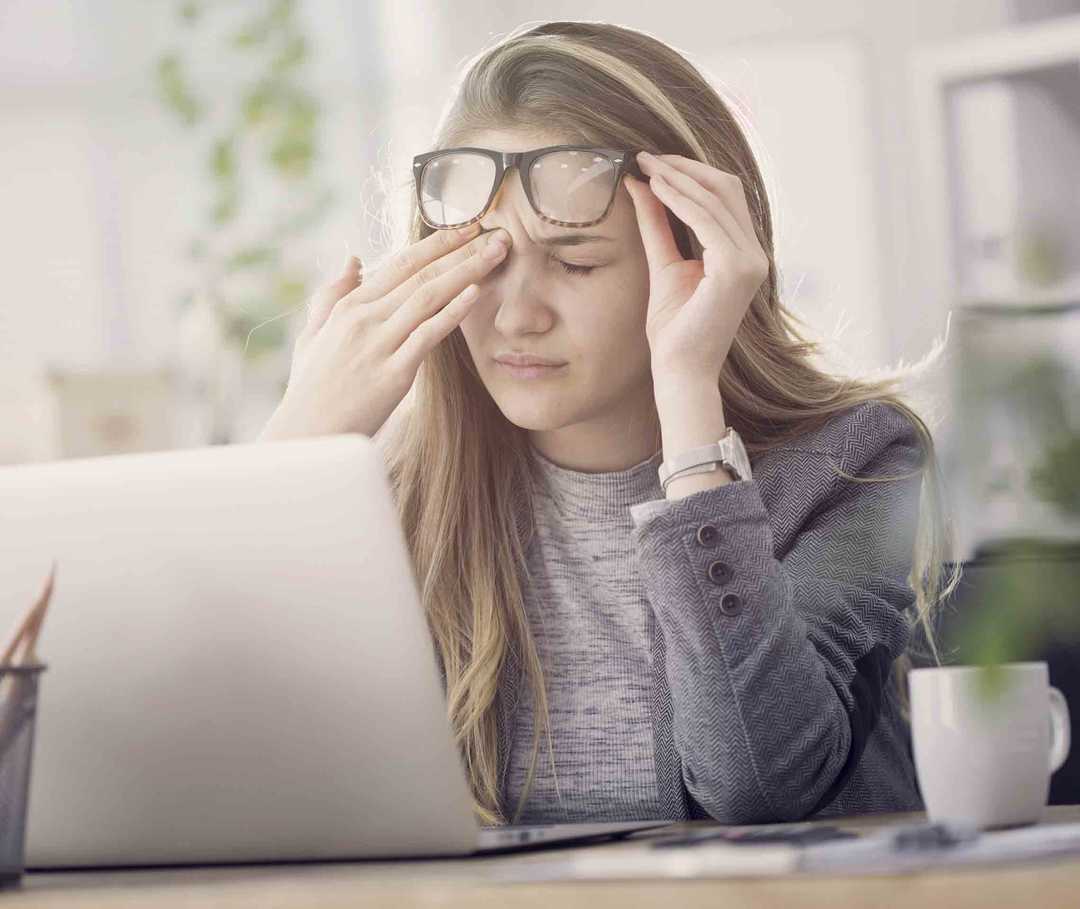 Frau mit schmerzverzogenem Gesicht sitzt mit Augenmigräne vor ihrem PC. Die Symptome können ein Flimmern vor den Augen oder ein Skotom sein.