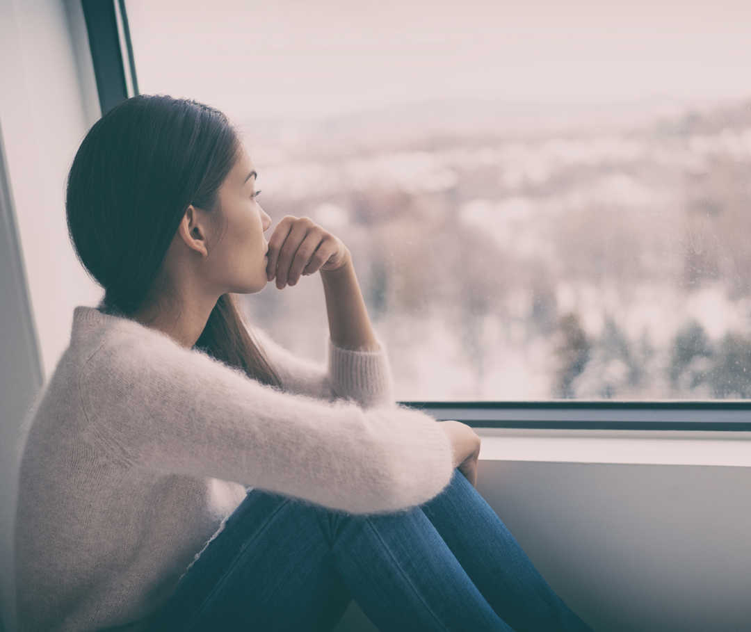 Frau mit Angst vor Migräne sitzt am Fenster und schaut hinaus.