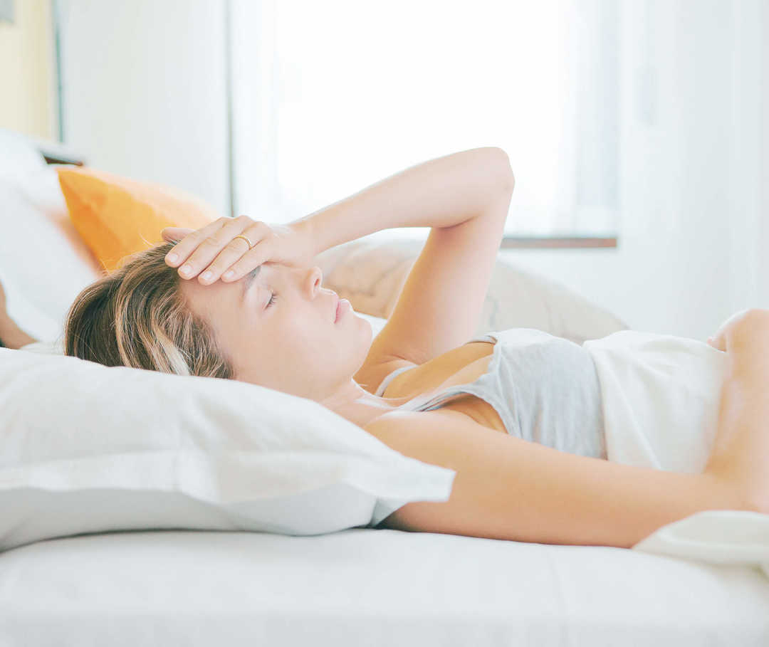 Frau mit Migräne und Übelkeit liegt im Bett und fragt sich, was sie dagegen tun soll.