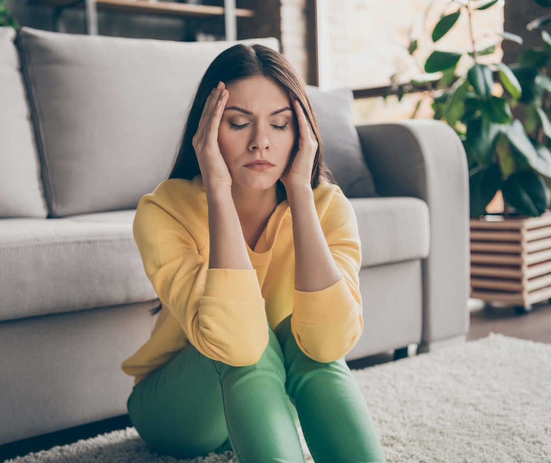 Frau sitzt vor der Couch und hat eine Migräne-Attacke durch Reizüberflutung. 