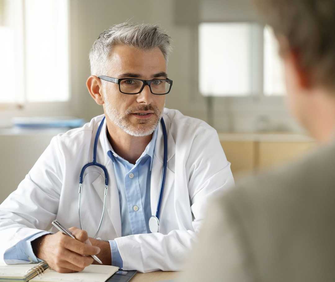 Ein Mann in weißem Kittel und mit einem Stethoskop um den Hals im Gespräch mit einem anderen Mann. 