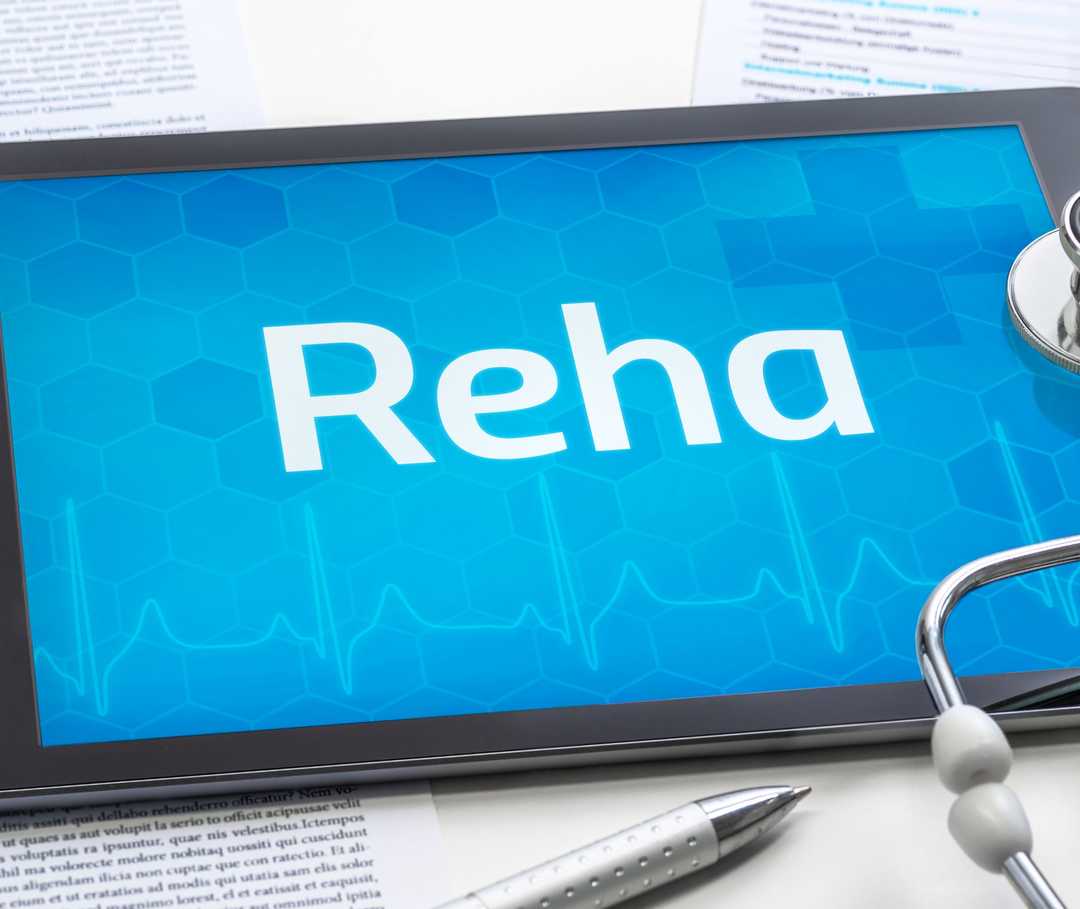 Reha oder Kur bei Migräne Auf dem Bildschirm eines Tablet-PCs ist das Wort Reha zu lesen. Daneben liegen ein Stethoskop, ein Stift und eine Blisterpackung mit Tabletten.