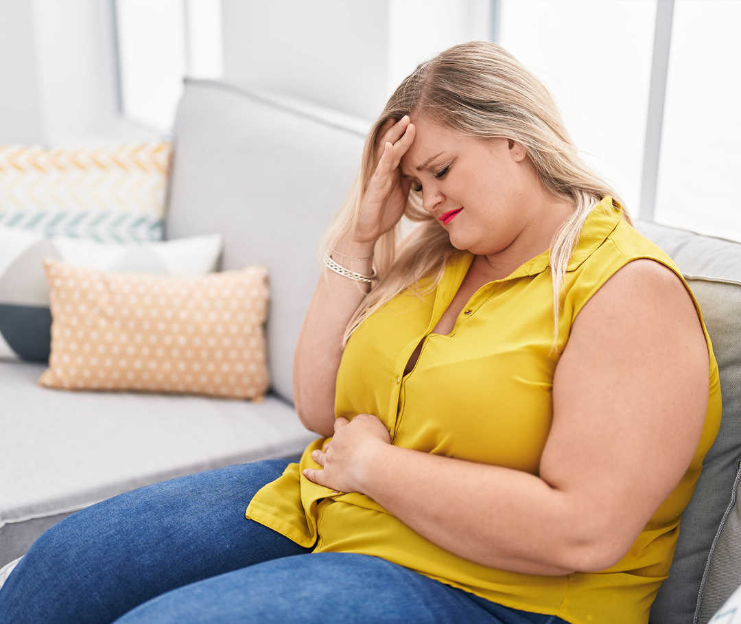Migräne und Darm Eine junge Frau sitzt auf einem Sofa und hält sich mit einer Hand den Kopf, mit der anderen den Bauch.