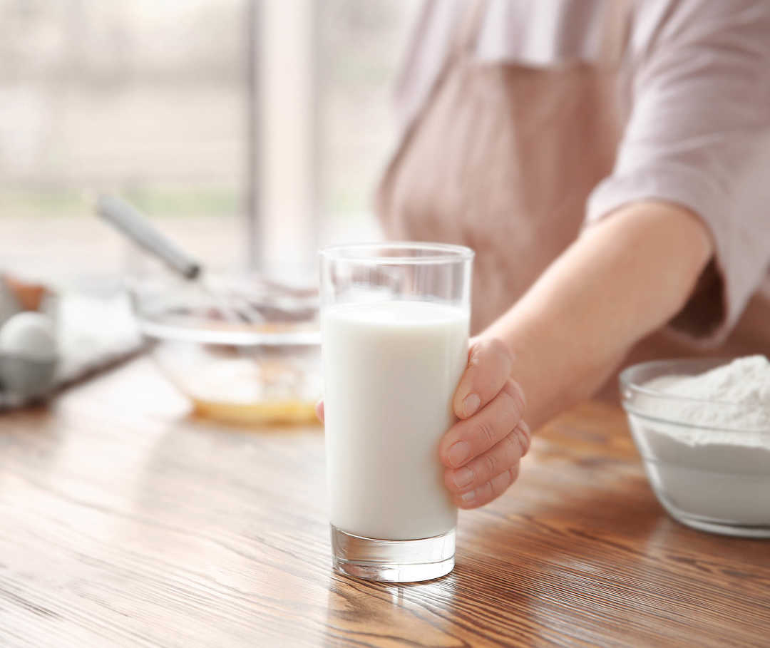 Eine Person greift nach einem Glas Milch auf dem Tisch, daneben stehen Backzutaten.