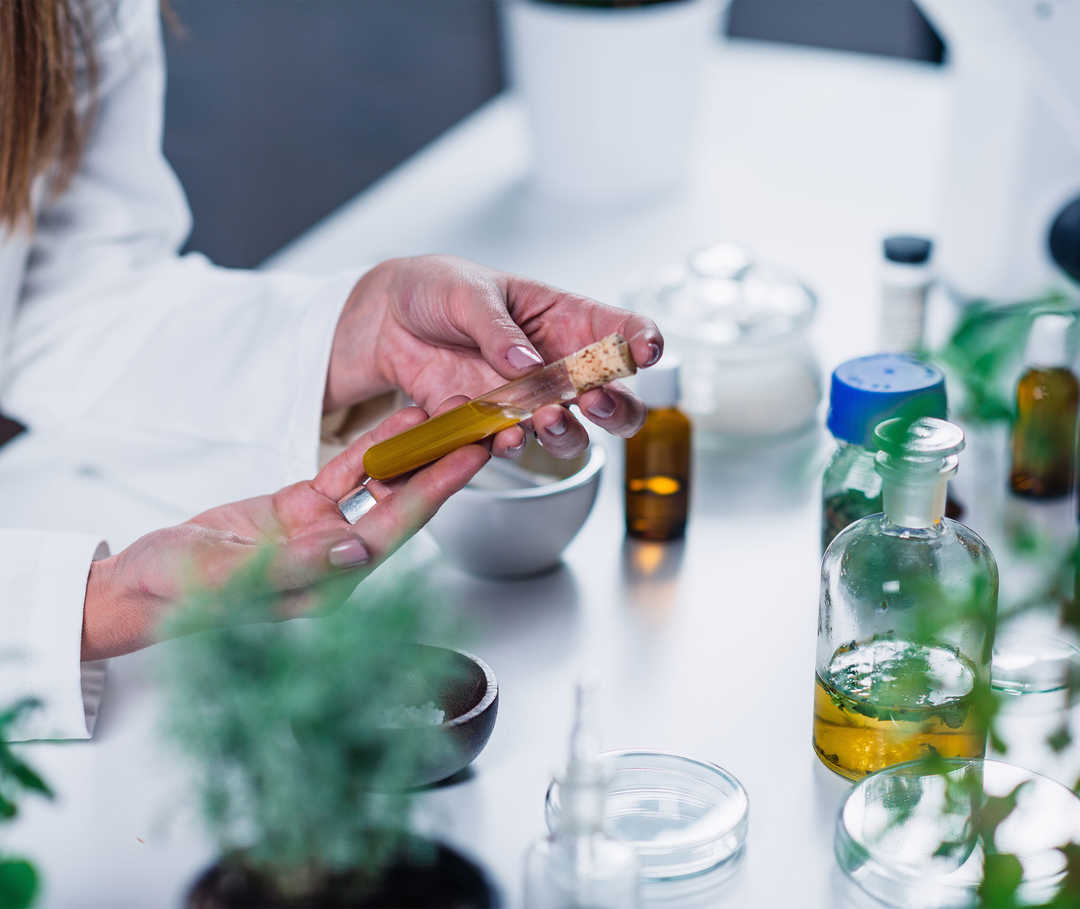 Eine Person in einem weißen Kittel hält ein Glasröhrchen mit einer braunen Flüssigkeit. 