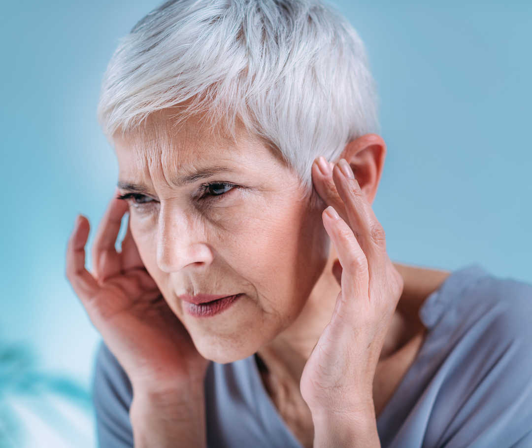 Migräne und Tinnitus Eine Frau mit kurzen grauen Haaren drückt mit ihren Fingern auf beide Ohren. Ihr Gesichtsausdruck ist angespannt.