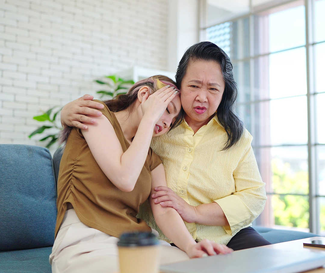 Eine Frau hält eine jüngere Frau im Arm, die sich die Hand auf die Schläfe legt.